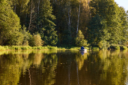 A lone fisherman in a boat on a peaceful lake, surrounded by dense green forest and reflected trees. Perfect for nature, outdoor activities, and fishing themesの写真素材