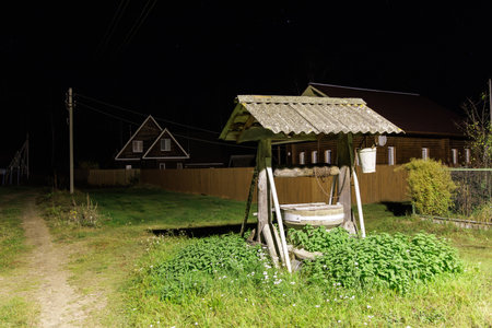 Nighttime view of a village featuring an old well with a wooden roof, illuminated by a streetlight. Traditional wooden houses with white-framed windows are visible in the backgroundの写真素材