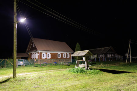 Nighttime view of a village featuring an old well with a wooden roof, illuminated by a streetlight. Traditional wooden houses with white-framed windows are visible in the backgroundの写真素材