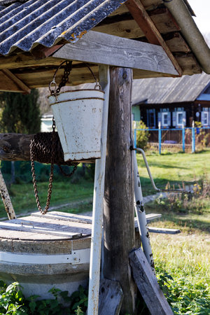 A detailed view of a rustic village well featuring a weathered metal bucket, a wooden beam, and a hand crank mechanism. The background includes a blurred view of a house and gardenの写真素材
