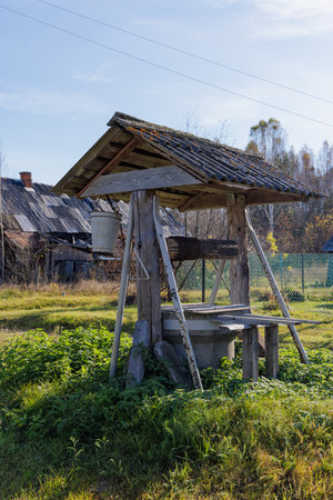 A serene morning scene featuring an old wooden well in a rural village, surrounded by traditional houses and lush greenery under a clear blue skyの写真素材