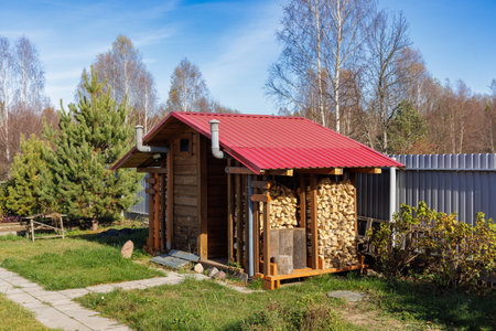 A traditional wooden outhouse with a red roof, located in a rural area, surrounded by greenery and a fenceの写真素材