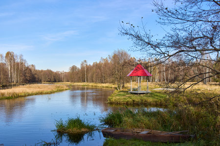 A serene lakeside scene featuring a gazebo with a red roof, surrounded by autumn trees and a wooden boat on the grassy shoreの写真素材