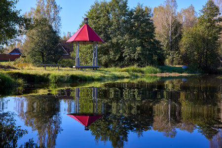A peaceful scene of a red-roofed gazebo reflected in still water, surrounded by lush greenery under a blue skyの写真素材