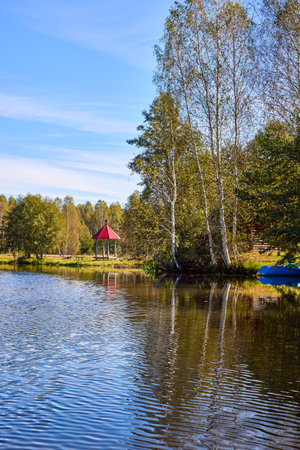 A peaceful scene of a red-roofed gazebo reflected in still water, surrounded by lush greenery under a blue skyの写真素材