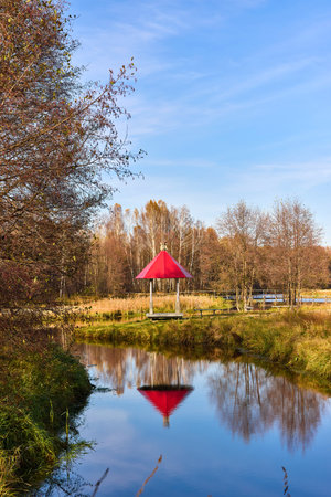 A peaceful scene of a red-roofed gazebo reflected in still water, surrounded by lush greenery under a blue skyの写真素材