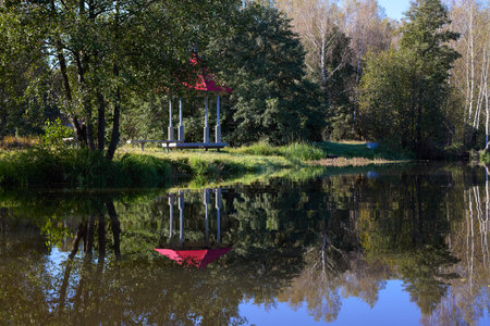 A peaceful scene of a red-roofed gazebo reflected in still water, surrounded by lush greenery under a blue skyの写真素材