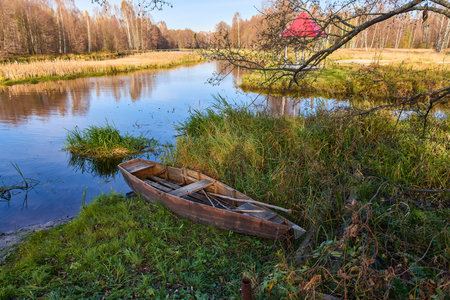A serene lakeside scene featuring a gazebo with a red roof, surrounded by autumn trees and a wooden boat on the grassy shoreの写真素材