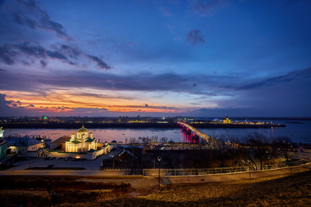 A mesmerizing sunset over the Annunciation Monastery, Strelka in Nizhny Novgorod, featuring Kanavinsky Bridge and the illuminated Alexander Nevsky Cathedral. The frozen river and glowing city lights create a stunning winter viewの写真素材