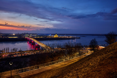 A mesmerizing sunset over Strelka in Nizhny Novgorod, featuring Kanavinsky Bridge and the illuminated Alexander Nevsky Cathedral. The frozen river and glowing city lights create a stunning winter viewの写真素材