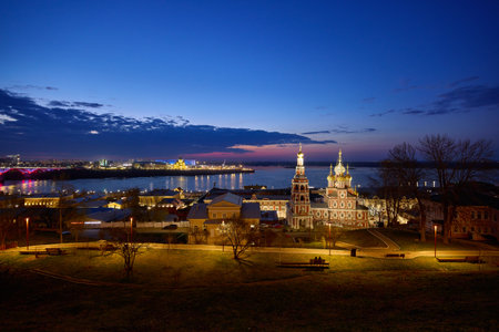 A captivating nighttime view of Strelka in Nizhny Novgorod, with the Oka River reflecting the glowing lights of Rozhdestvenskaya Church and Alexander Nevsky Cathedral under a twilight skyの写真素材
