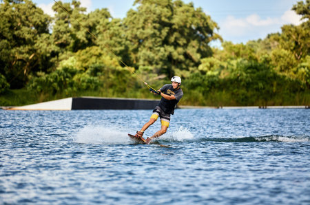 Young man wakeboarding on a lake, wearing helmet and gloves, riding smoothly with a splash trailの写真素材