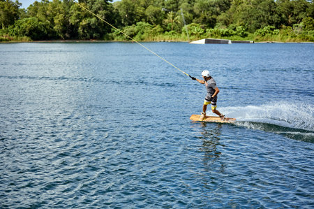 Young man wakeboarding on a lake, wearing helmet and gloves, riding smoothly with a splash trailの写真素材