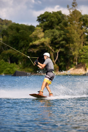 Young man wakeboarding on a lake, wearing helmet and gloves, riding smoothly with a splash trailの写真素材