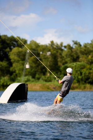Young man wakeboarding on a lake, wearing helmet and gloves, riding smoothly with a splash trailの写真素材