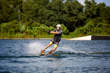 Young man wakeboarding on a lake, wearing helmet and gloves, riding smoothly with a splash trailの写真素材