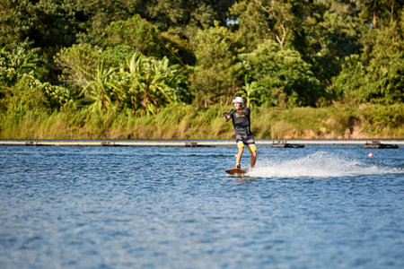 Young man wakeboarding on a lake, wearing helmet and gloves, riding smoothly with a splash trailの写真素材
