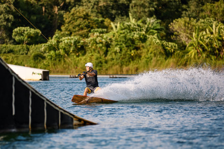 Young man wakeboarding on a lake, wearing helmet and gloves, riding smoothly with a splash trailの写真素材