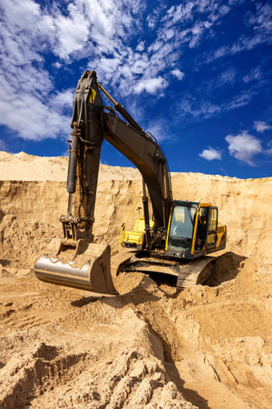 Large yellow excavator operating in a sandy quarry with dramatic blue sky and clouds in the backgroundの写真素材