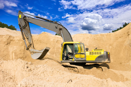 Large yellow excavator operating in a sandy quarry with dramatic blue sky and clouds in the backgroundの写真素材