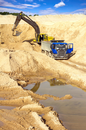 Yellow excavator loads sand into dump truck at a construction quarry under a vibrant blue skyの写真素材