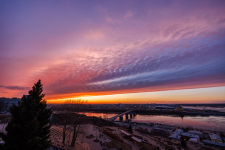 A mesmerizing sunset over Strelka in Nizhny Novgorod, featuring Kanavinsky Bridge and the illuminated Alexander Nevsky Cathedral. The frozen river and glowing city lights create a stunning winter viewの写真素材