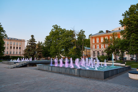 A vibrant fountain lights up Markin Square in Nizhny Novgorod at evening, surrounded by lush trees and historic buildings under a twilight skyの写真素材