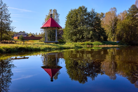 A peaceful scene of a red-roofed gazebo reflected in still water, surrounded by lush greenery under a blue skyの写真素材