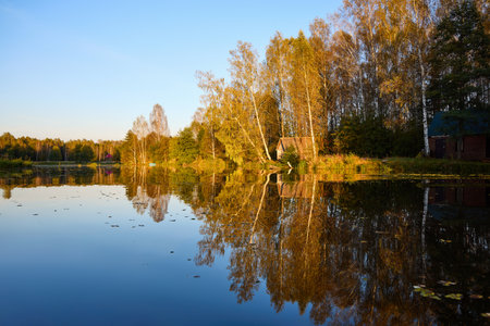 A serene riverside scene with a wooden house and lush trees, perfectly reflected in the calm waterの写真素材