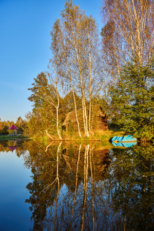 A serene riverside scene with a wooden house and lush trees, perfectly reflected in the calm waterの写真素材