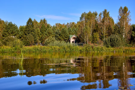 A serene riverside scene with a wooden house and lush trees, perfectly reflected in the calm waterの写真素材