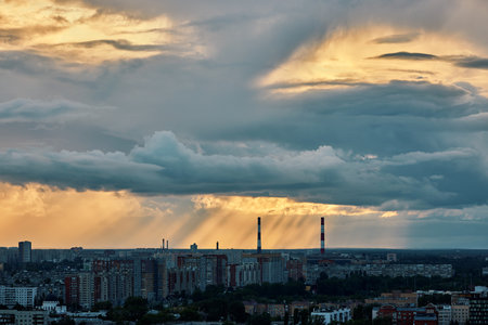 Golden rays of sunset pierce through dramatic clouds over a cityscape with residential buildings and chimneysの写真素材