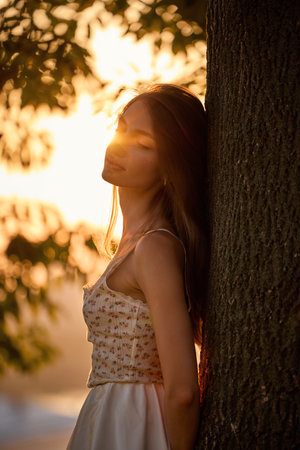 Slavic girl in floral lace dress stands near tree in golden sunlight, serene and stylishの写真素材