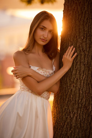 Slavic girl in floral lace dress stands near tree in golden sunlight, serene and stylishの写真素材
