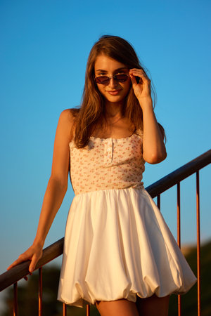 Slavic girl in floral white dress and sunglasses stands on railing in warm evening sunlightの写真素材