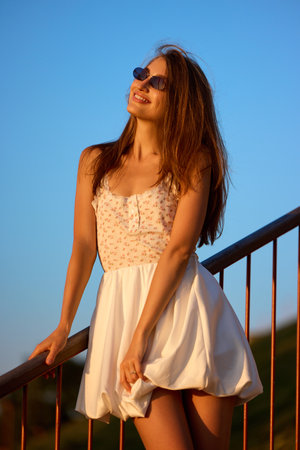 Slavic girl in floral white dress and sunglasses stands on railing in warm evening sunlightの写真素材