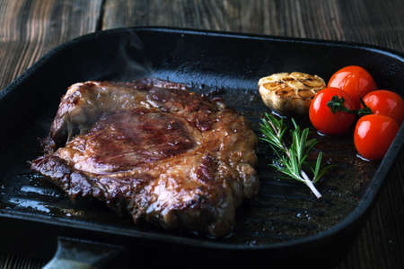 Grilled ribeye steak on a grill pan with rosemary and garlic on a wooden background