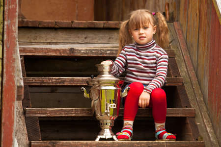 Funny lovely little girl posing sitting near the Russian Samovar on the porch of the farmhouse.の写真素材