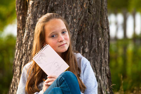 Teen-girl shows notebook while sitting in the park  looks into the camera の写真素材