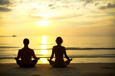 Yoga practice, a young couple sitting on the beach of the sea in the lotus position at sunset の写真素材