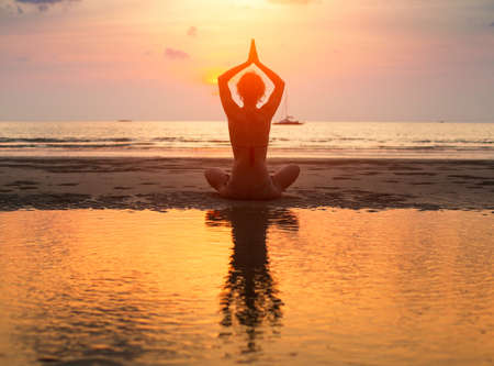 Yoga woman sitting in lotus pose on the beach during sunset with reflection in water.の写真素材