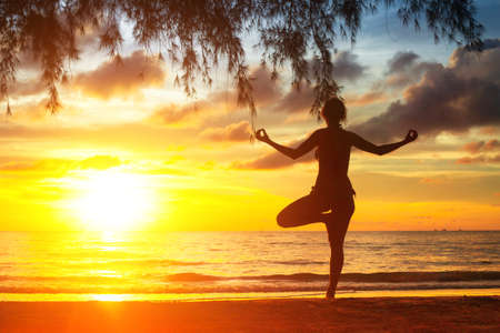 Silhouette of young woman practicing yoga on the sea beach at sunset.の写真素材