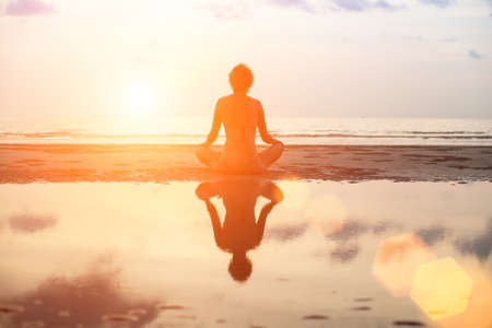 Yoga woman sitting in lotus pose on the beach during sunset, with reflection in water.の写真素材