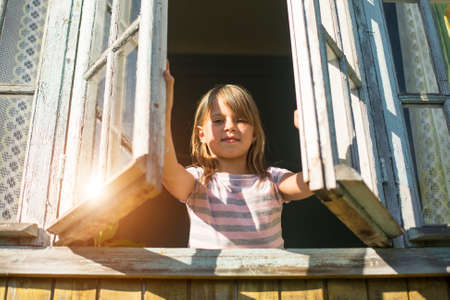 Beautiful little girl looks out the window rural houseの写真素材