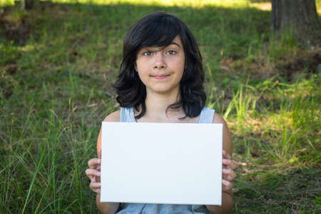 Lovely teen-girl holding clean white sheet paper, in the park (banner for your message)の写真素材