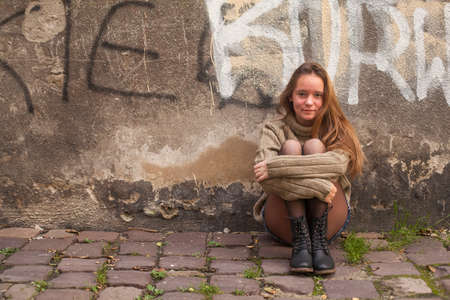 Pretty young girl sitting on the pavement near a stone wall of a house.の写真素材