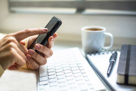 Businesswoman using mobile phone, cup of coffe, computer keyboard and notepad in the background.