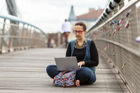 Young woman with laptop sitting on a pedestrian bridge in an old European city.