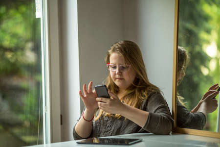 Teenage girl at glasses with smartphone sitting near the window.の写真素材