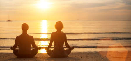 Man and woman yoga silhouettes meditating on Sea coast during the sunset.の写真素材